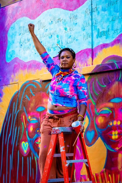 站在涂鸦墙旁边的一位女士，身穿蓝色和粉色花朵纽扣衬衫和棕色裤子